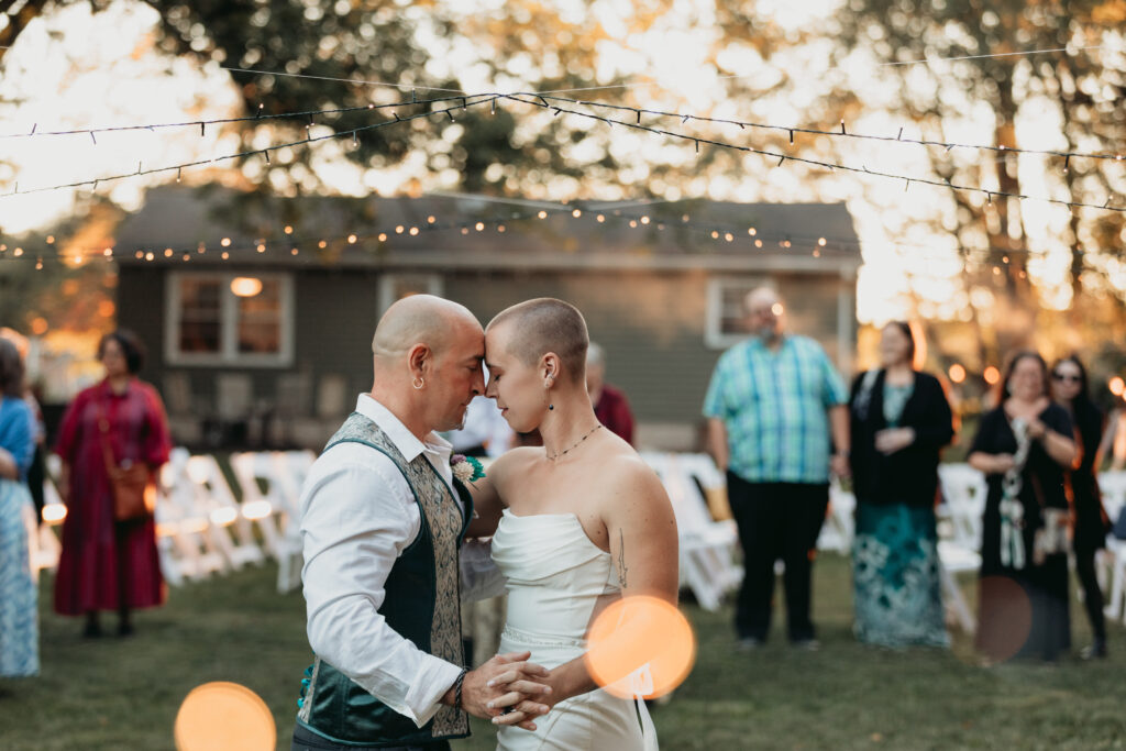 Couple sharing a quiet first dance during an outdoor wedding reception, surrounded by guests and string lights at sunset.