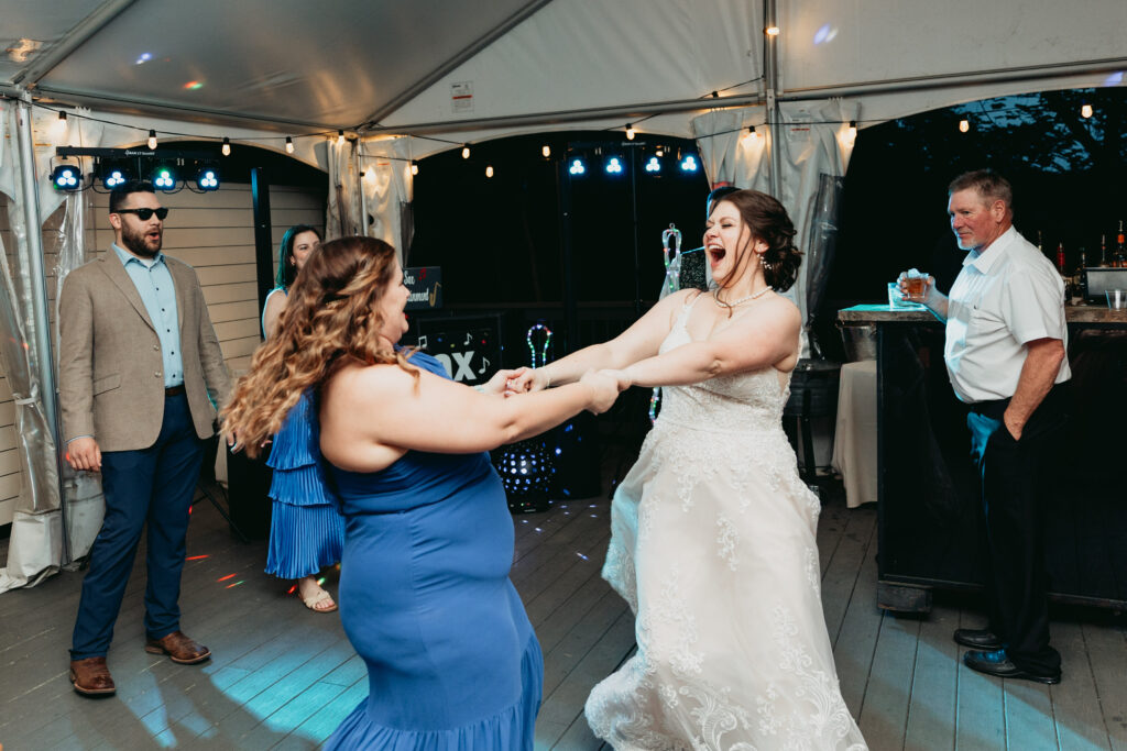 Bride laughing and dancing with a guest during a wedding reception inside a tent, surrounded by music and celebration.